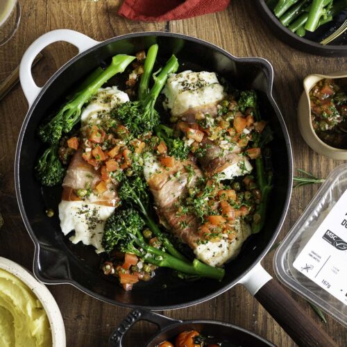 Kabeljauwhaasje in parmaham met sauce vierge en parmezaanpuree bovenaanzicht op tafel met gepofte tomaten en gestoomde bimi erbij