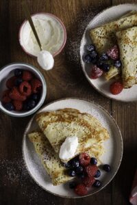 Gistpannenkoeken op 2 borden met slagroom, frambozen en bosbessen, en poedersuiker